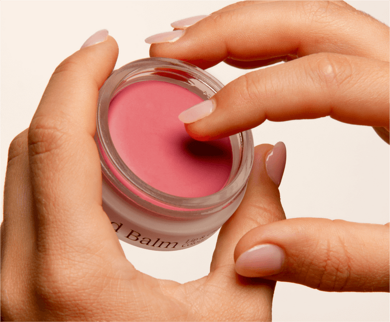 Fingers applying pink tinted balm from a small jar.